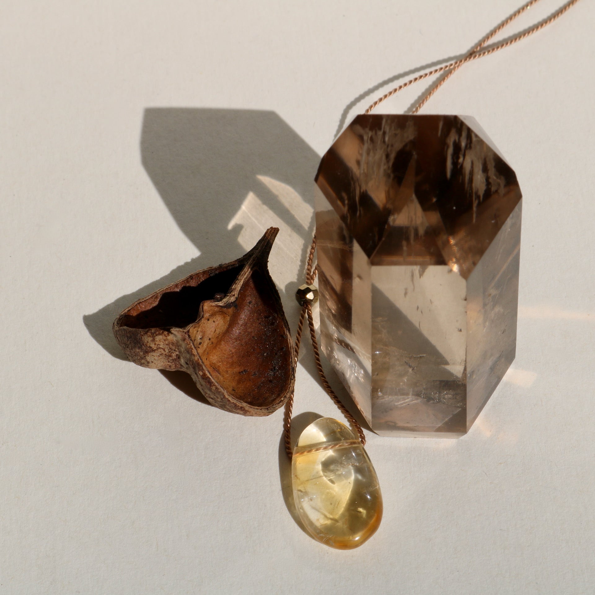 Smooth Citrine pebble-like stone on silk necklace displayed with smoky quartz pillar and seed pod in the sunshine on neutral cream colored background