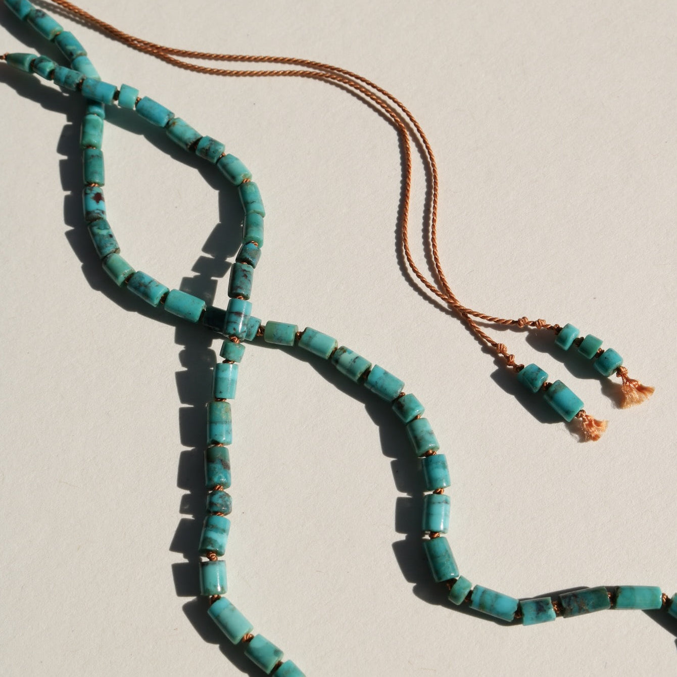 A turquoise beaded necklace with cylinder beads and a terracotta silk cord, laid out on a light beige background.