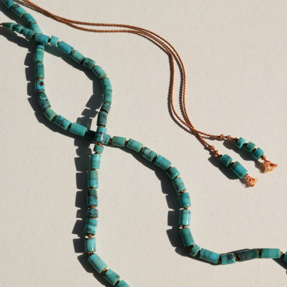 A turquoise beaded necklace with cylinder beads and a terracotta silk cord, laid out on a light beige background.